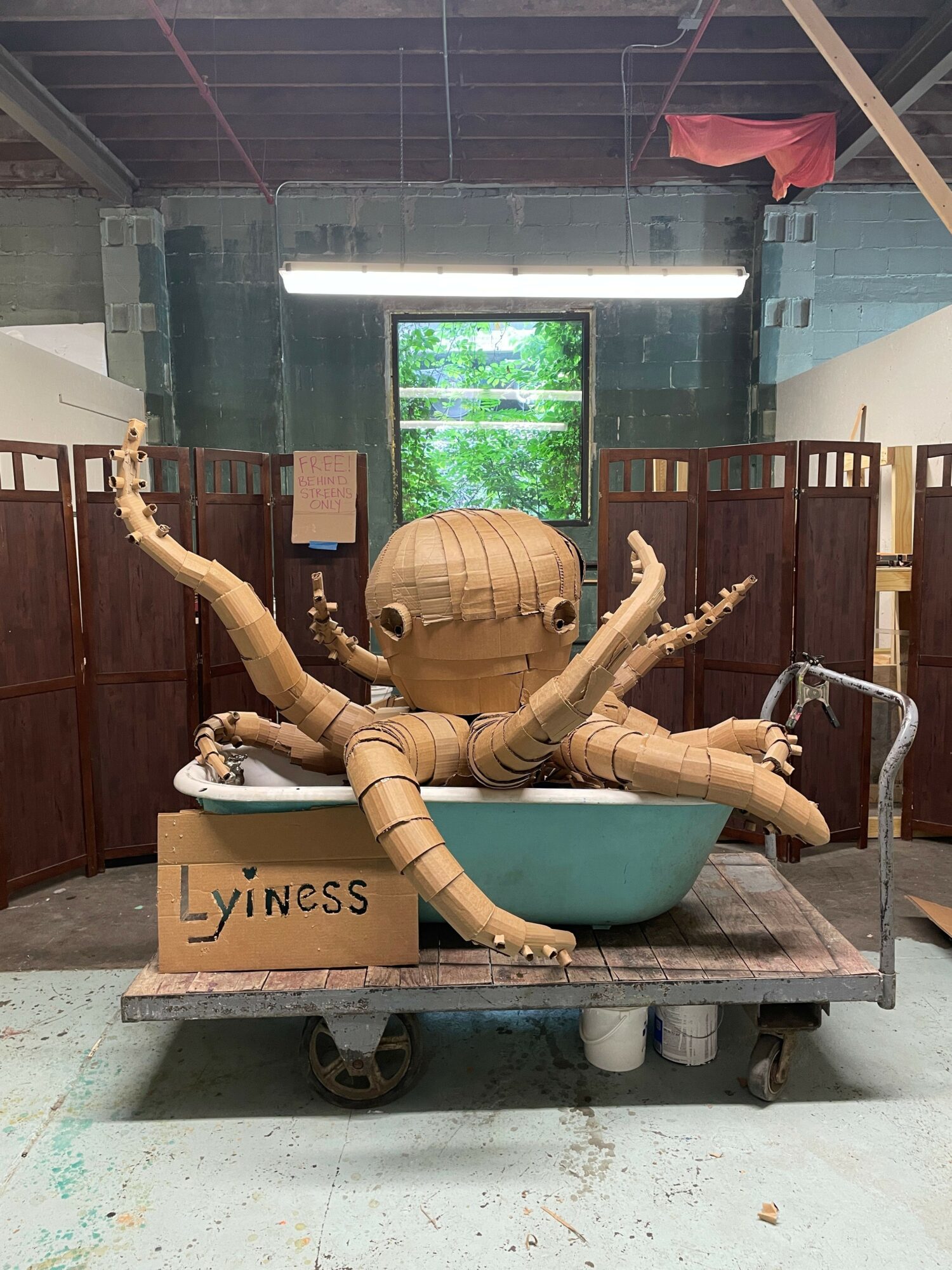 Large octopus sculpture with a round head and multiple arms, placed in a basin on a cart inside a building.