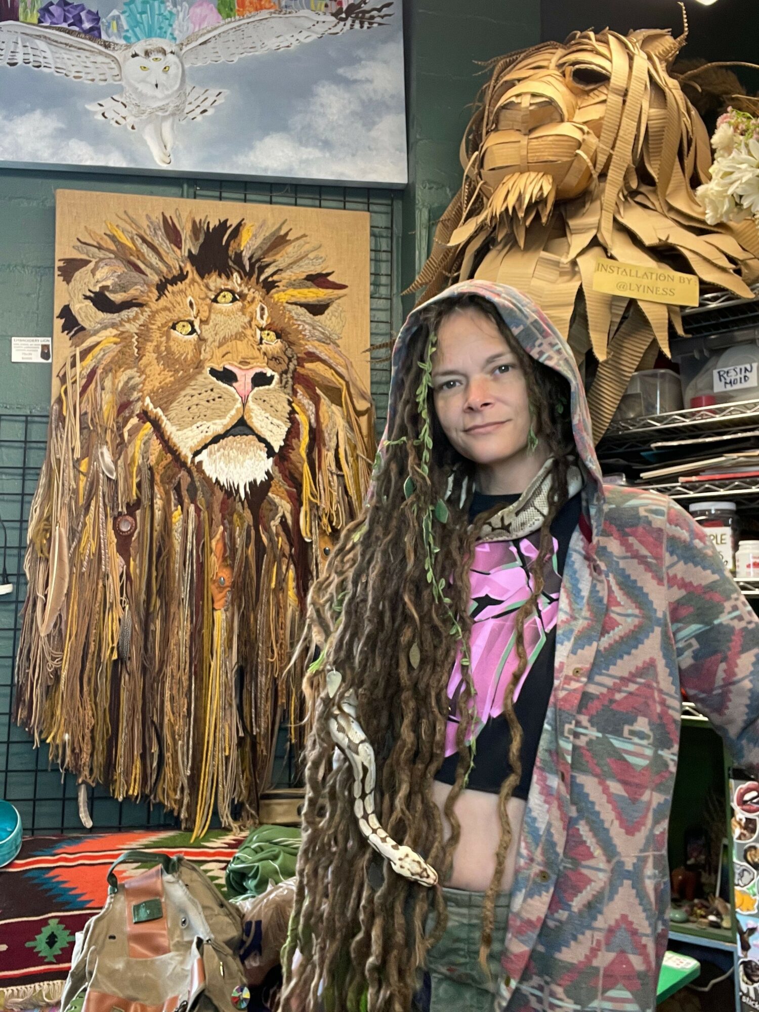 Girl with long dreadlocks wearing a patterned hoodie, standing next to a lion painting in an art store.