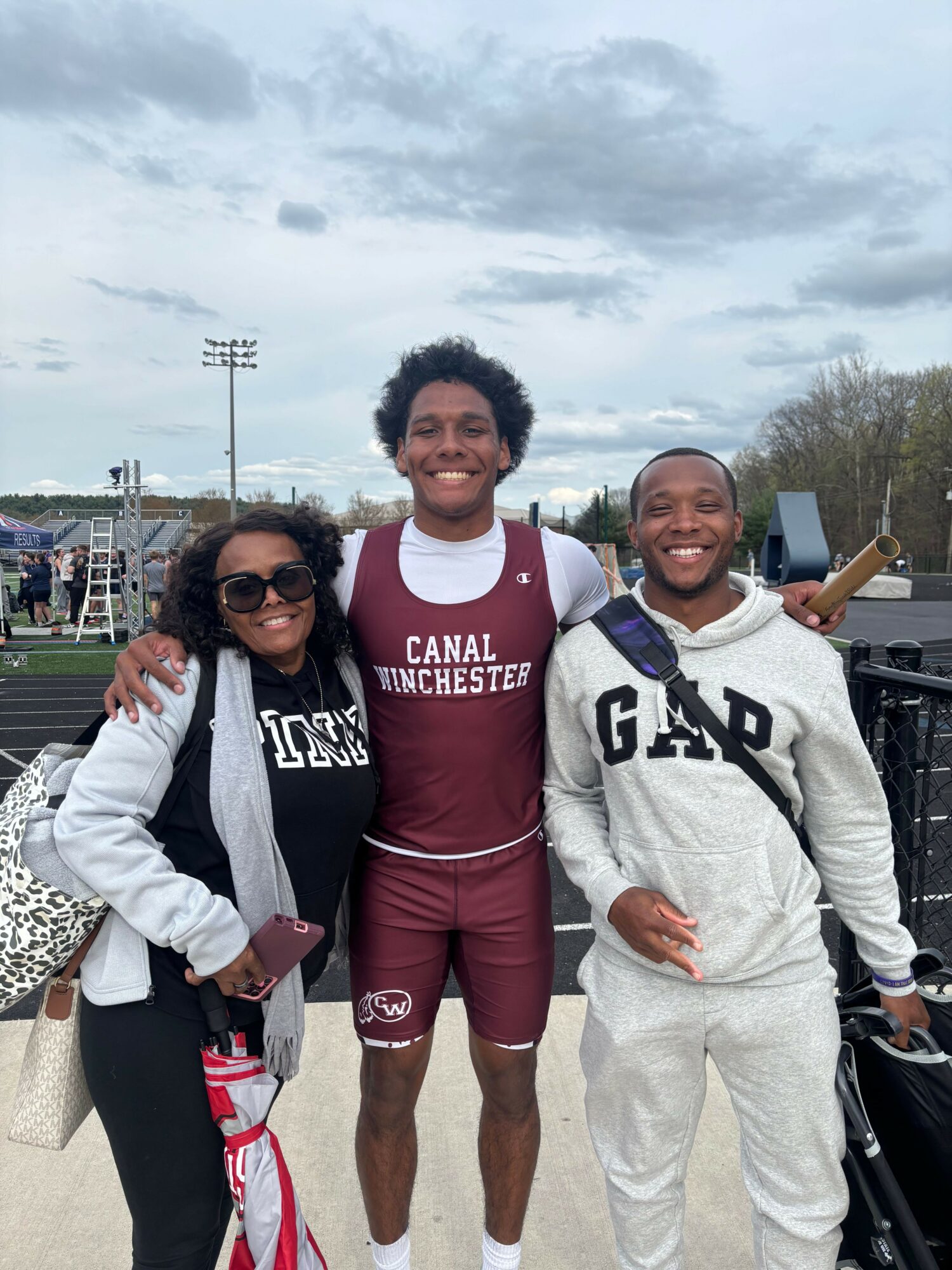 Three smiling people standing outdoors, one with arms around others, in a sports field setting.