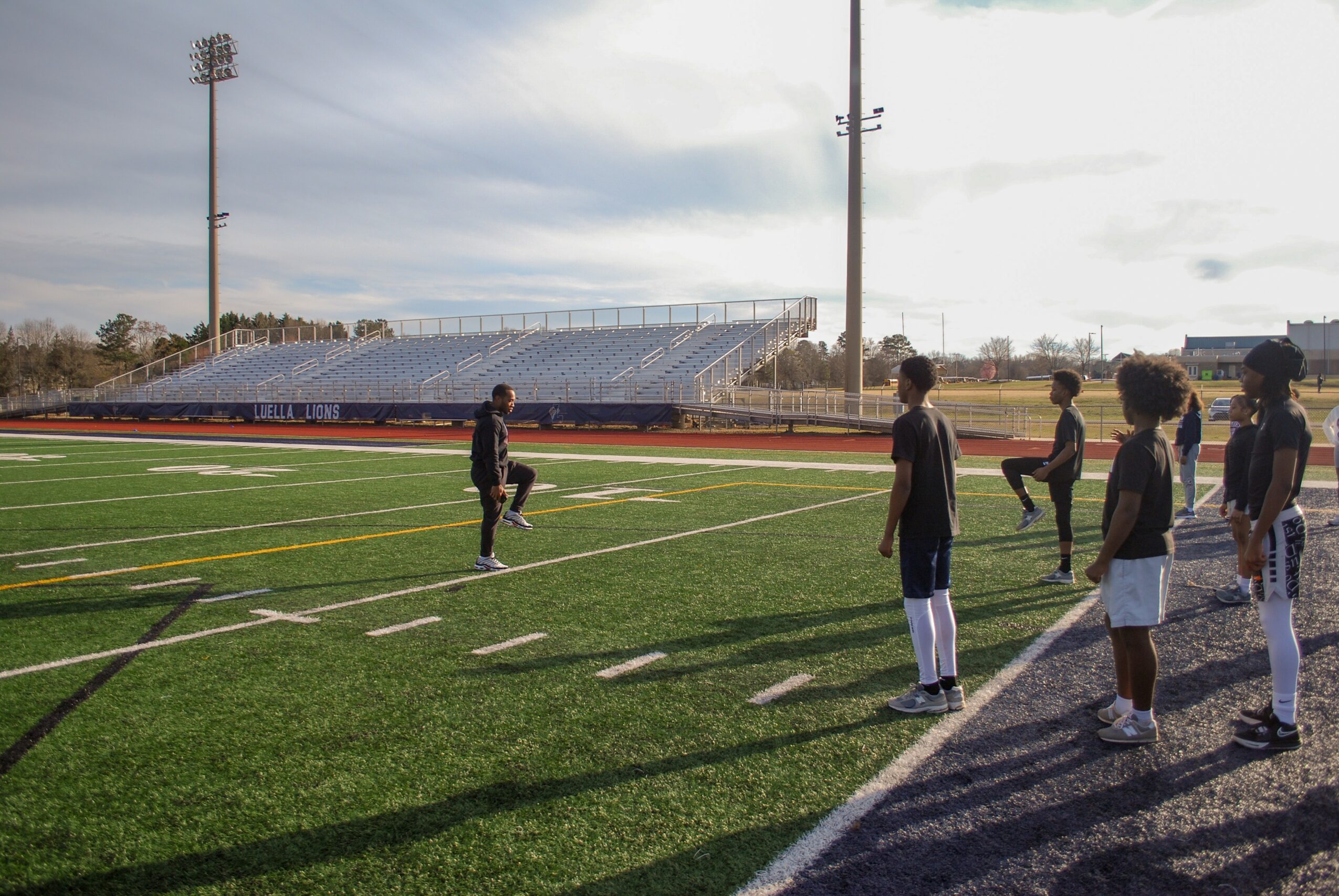 Group of people standing on a sports field with a stadium and sky in the background.