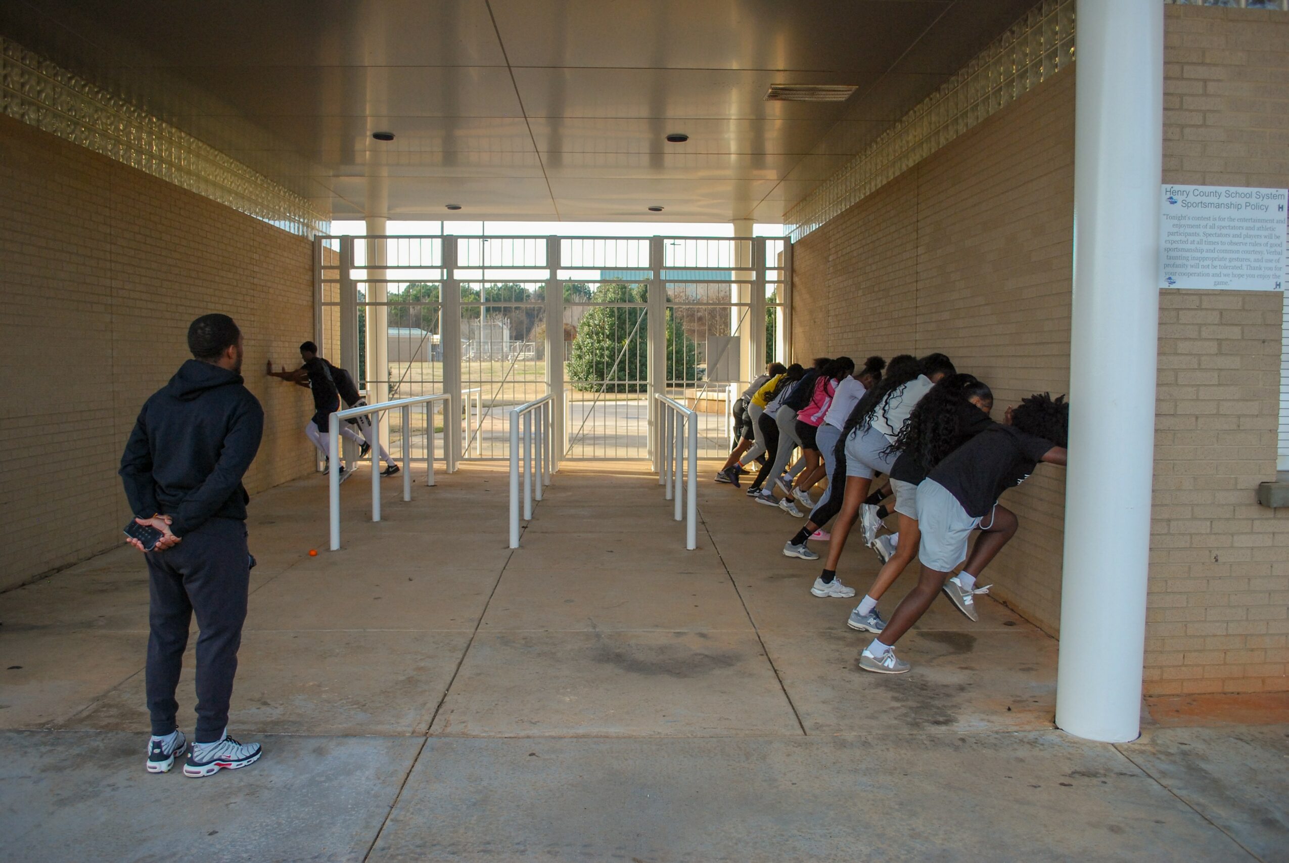 Group of students leaning forward against wall, facing away, with one person observing nearby in an open corridor.