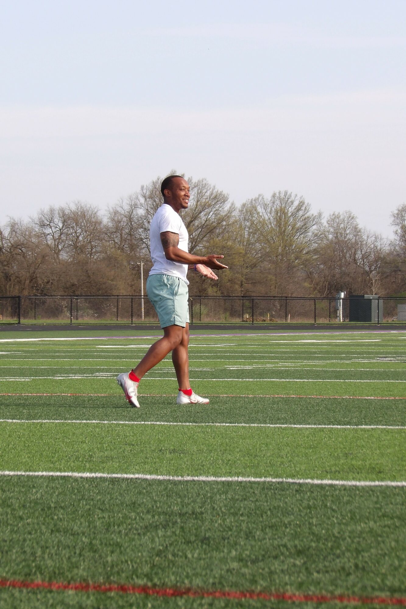 Person standing on a sports field with trees and a fence in the background.