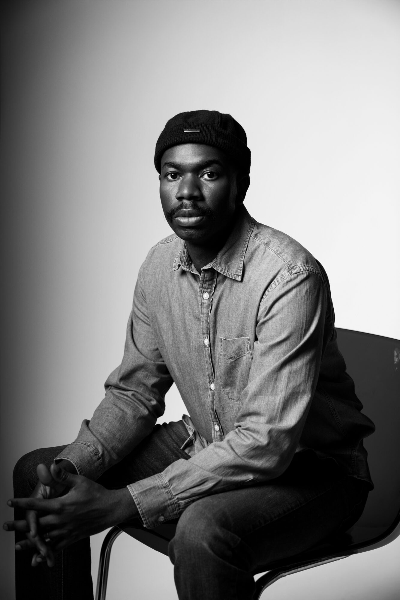 Young man sitting on a chair, wearing a denim shirt and a cap, looking at the camera.