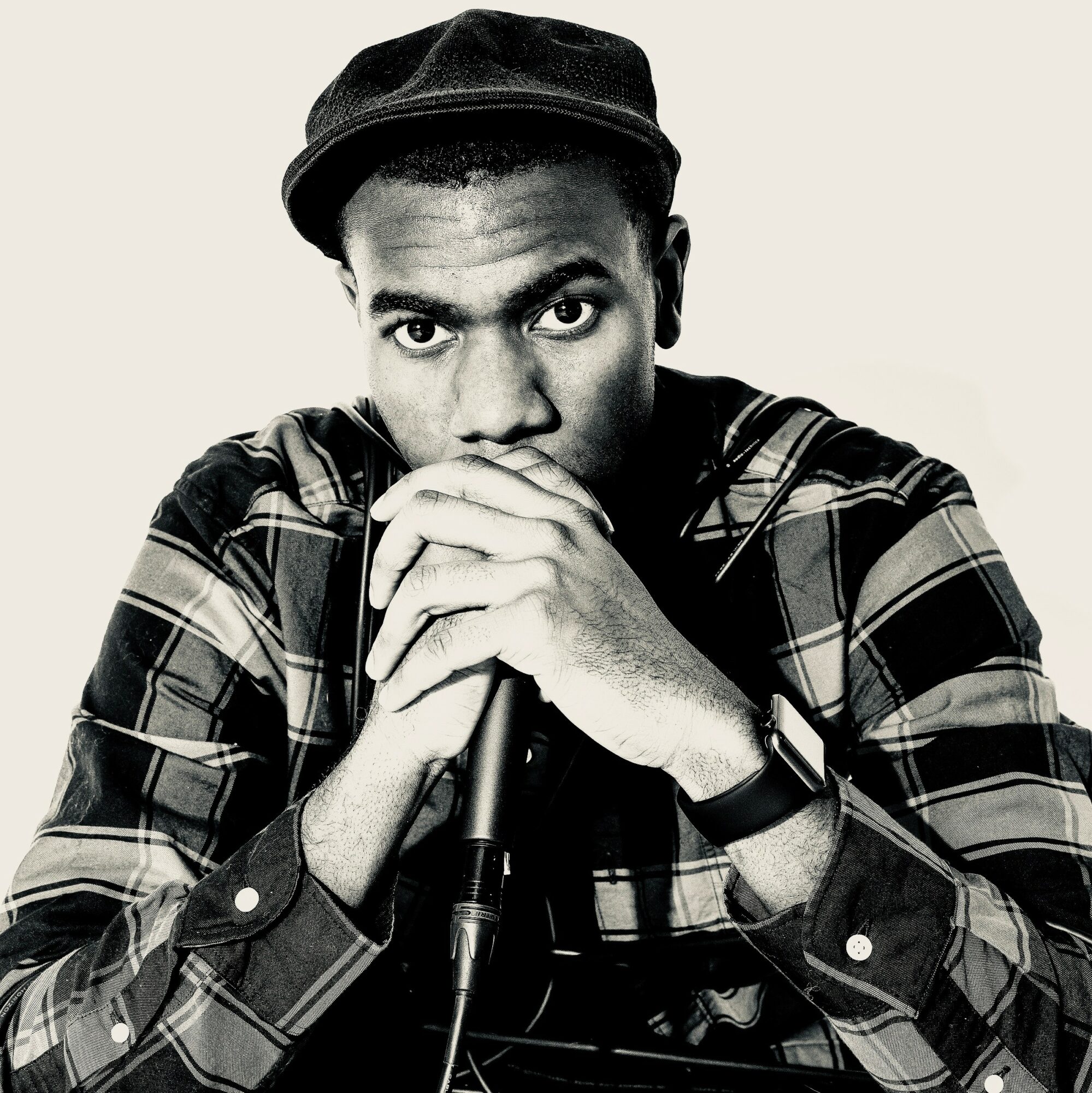 Young man wearing a cap and plaid shirt, holding a microphone with both hands, looking at the camera.