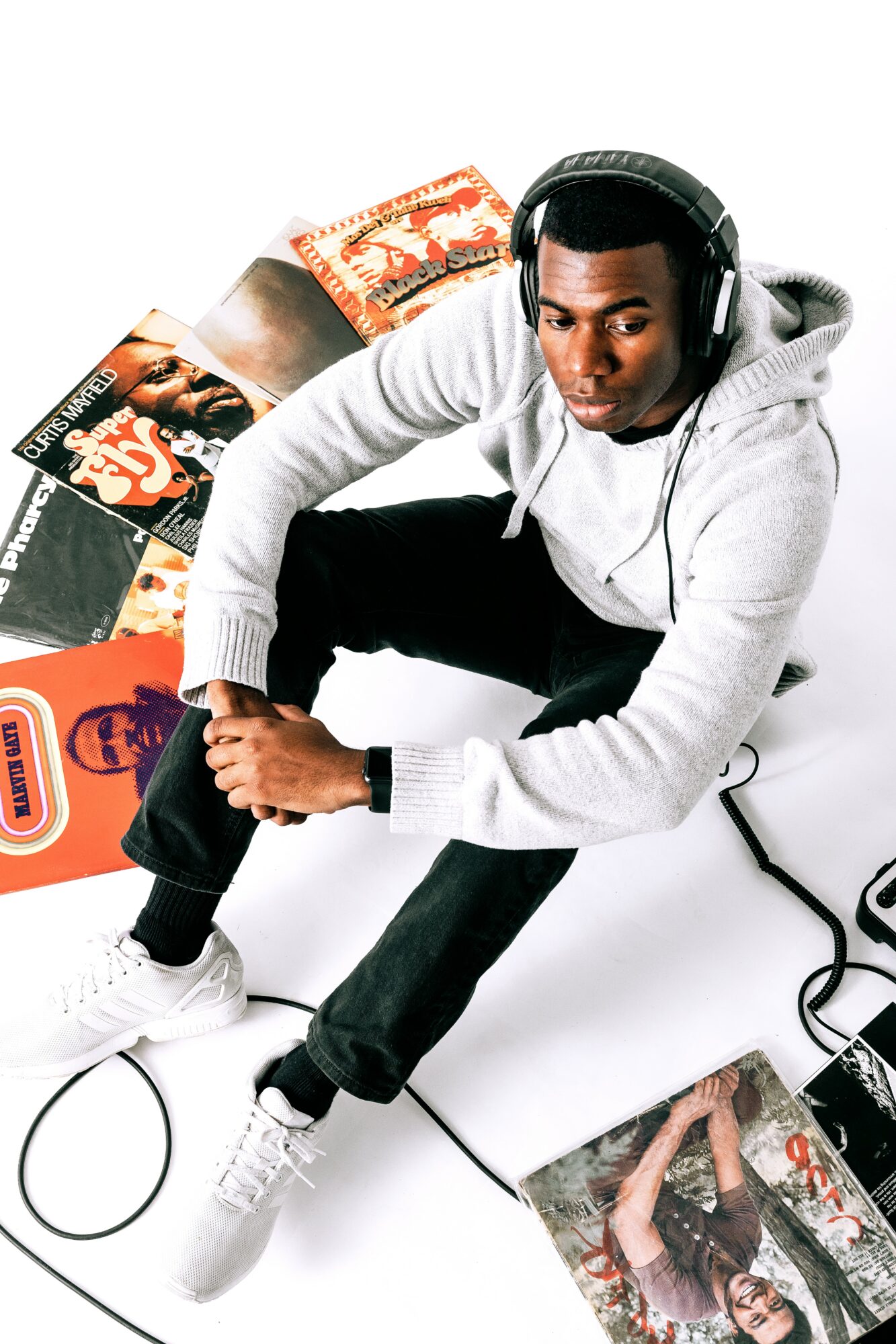 Young man wearing headphones, sitting with vinyl records and magazines around him, white background.