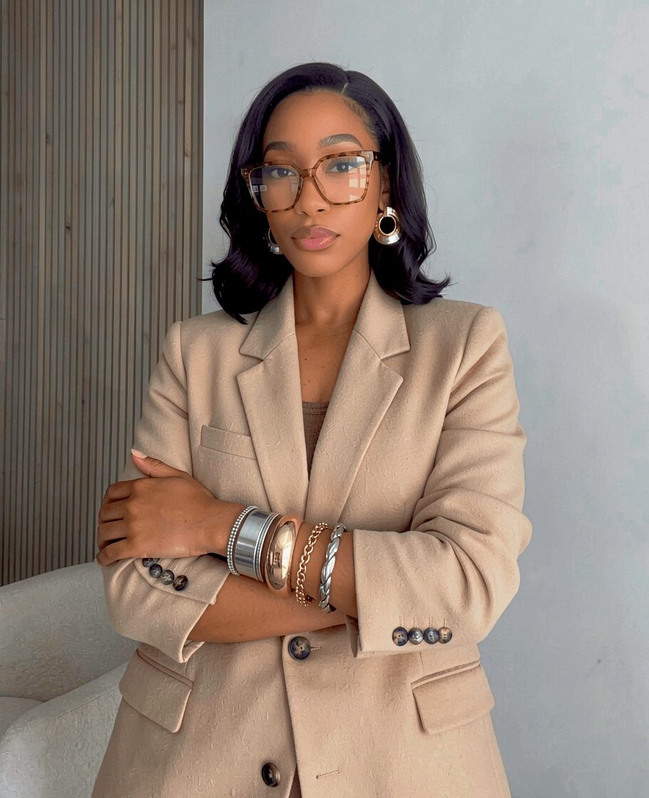 Woman with glasses and earrings wearing a beige blazer, standing with arms crossed against a plain background.
