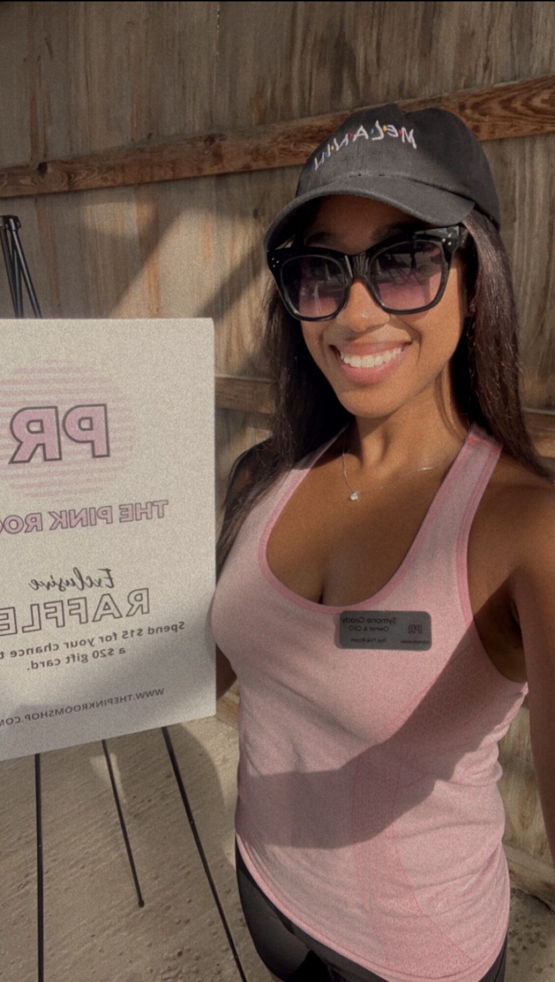Woman smiling wearing sunglasses, a cap, and a pink tank top, standing outdoors next to a sign.