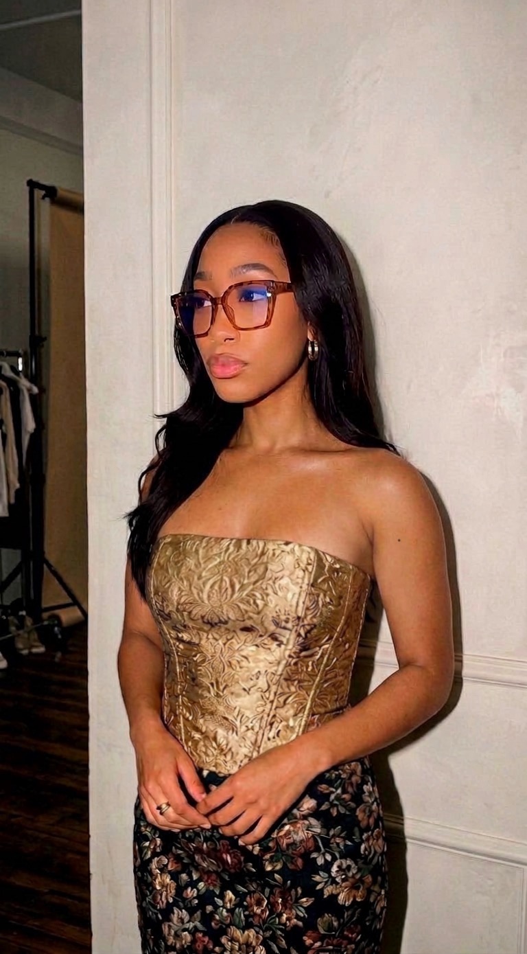 Young woman with long dark hair and glasses, wearing a strapless gold top and floral skirt, standing indoors.