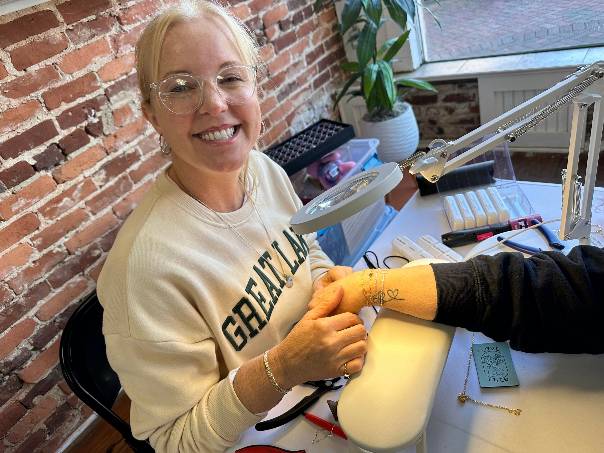 Woman with glasses smiling, receiving a manicure from a person wearing black sleeve, in a room with brick wall and plants.