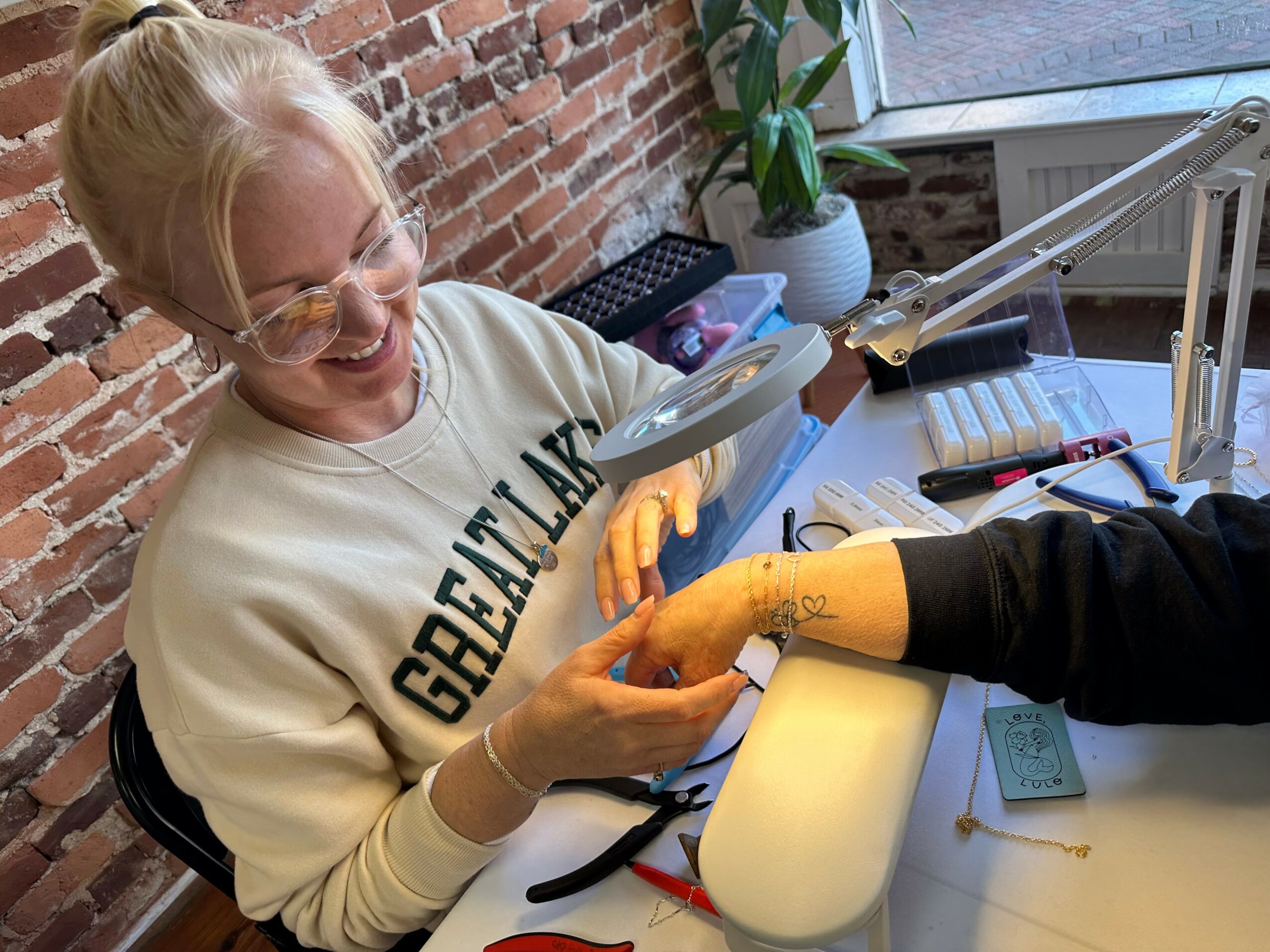 Person smiling and examining a hand held by another person in a bright workspace with brick wall and plants.