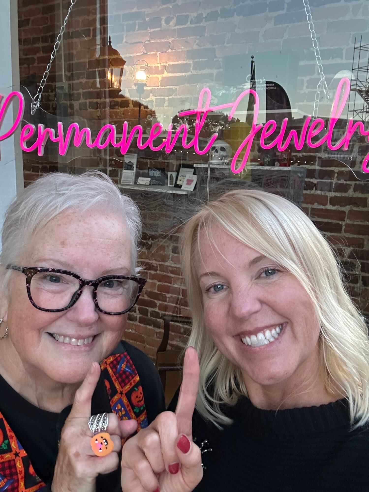 Two women smiling in front of a brick wall with a neon sign reading 'Permanent Jewelry'.