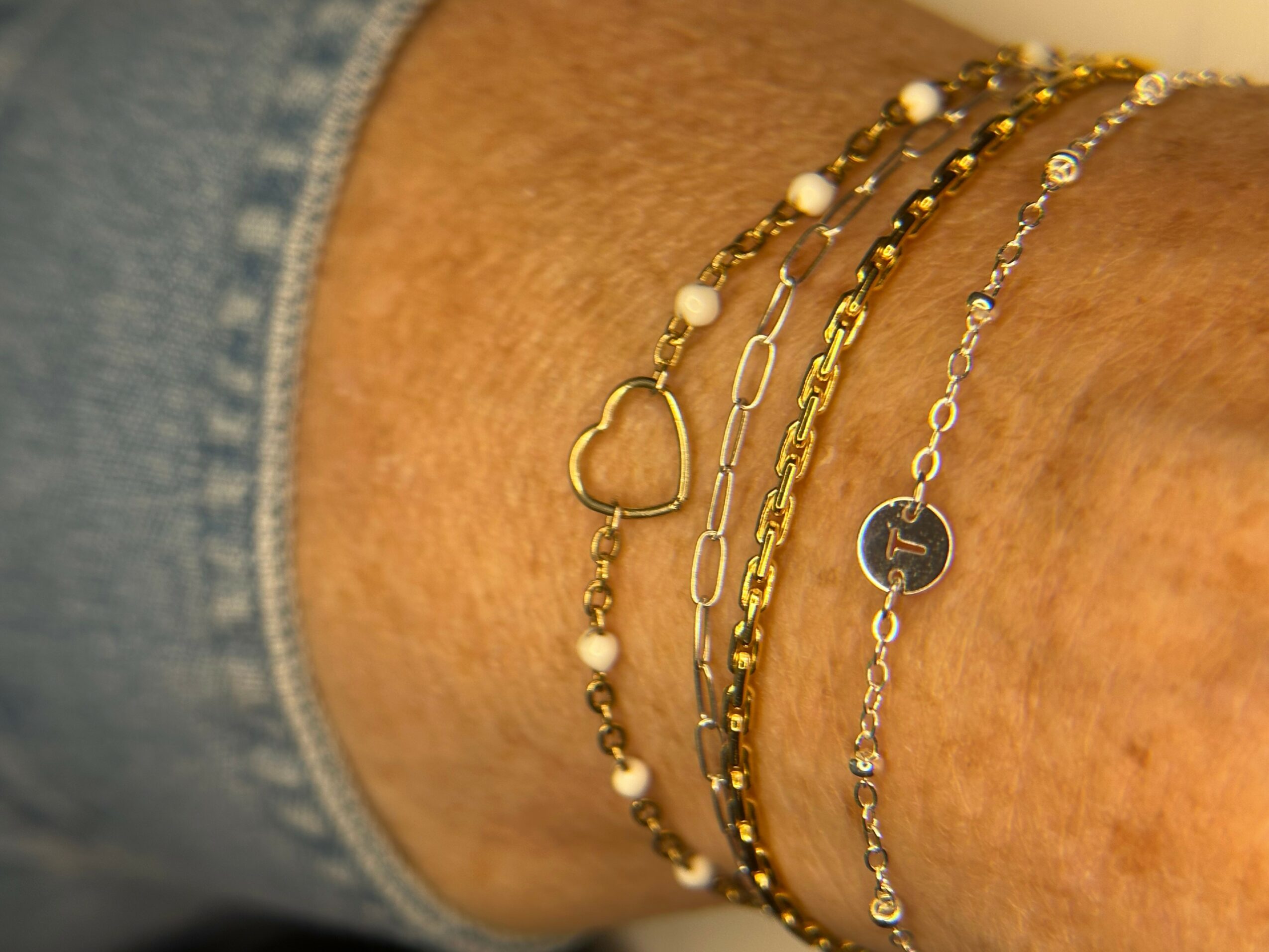 Four layered bracelets on a person's wrist, including a gold chain, a heart charm, and beaded designs.