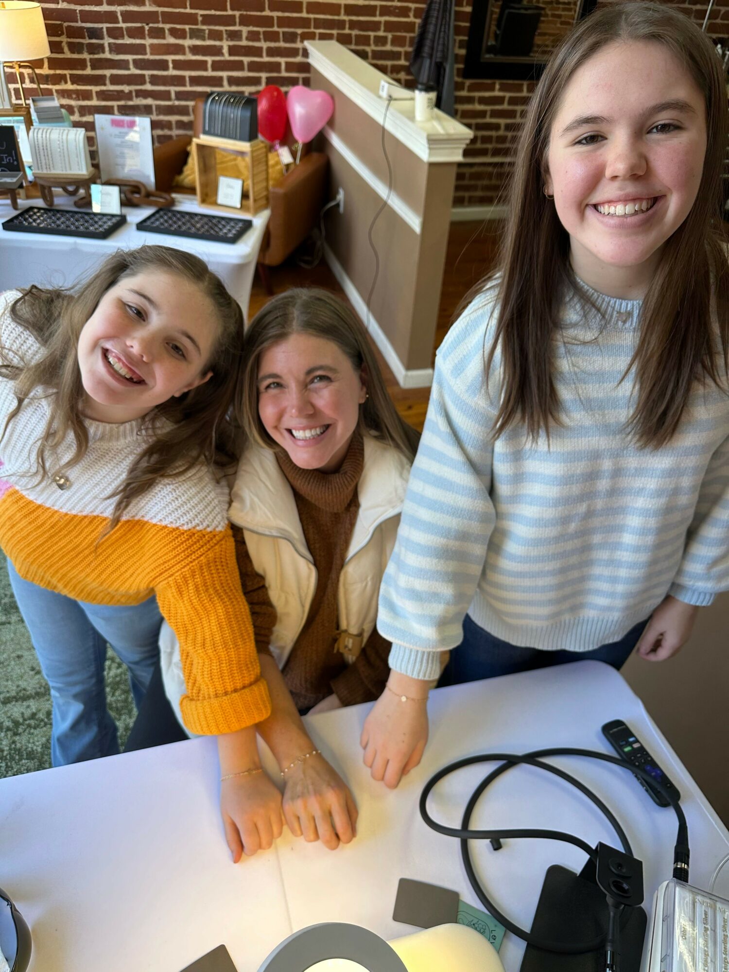 Three smiling girls gathered around a table, with one girl pointing at something on the table, in a room with a brick wall and office supplies.