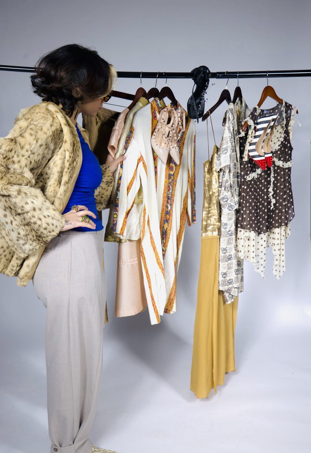 Woman looking at clothing rack with various dresses and pants in a room with white background.
