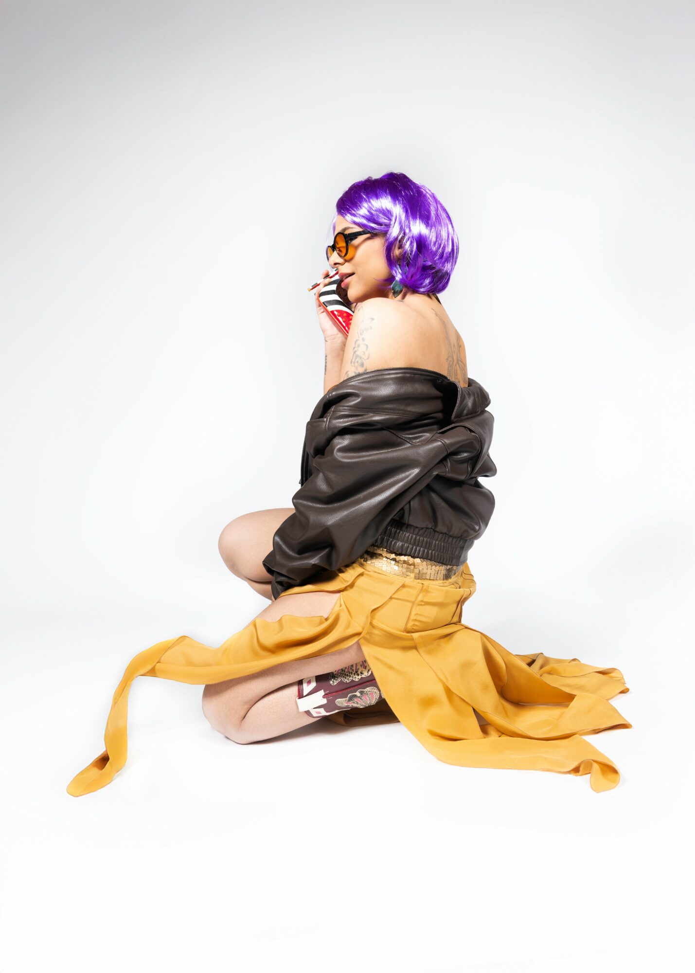 Person with purple hair and glasses kneeling on the floor, wearing a black jacket and yellow skirt, against a plain white background.