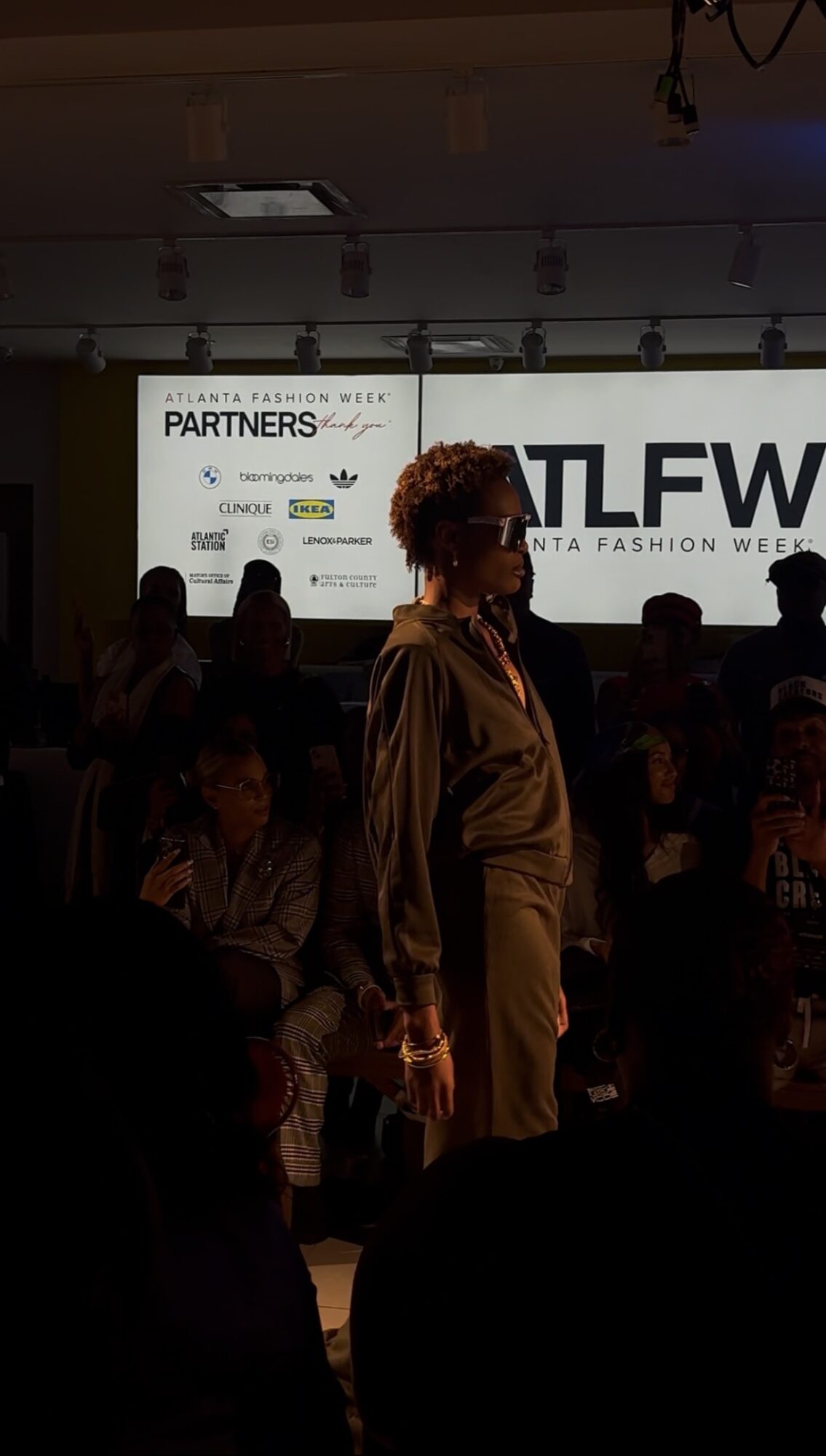 Person with curly hair standing in front of a large illuminated sign at a fashion event.