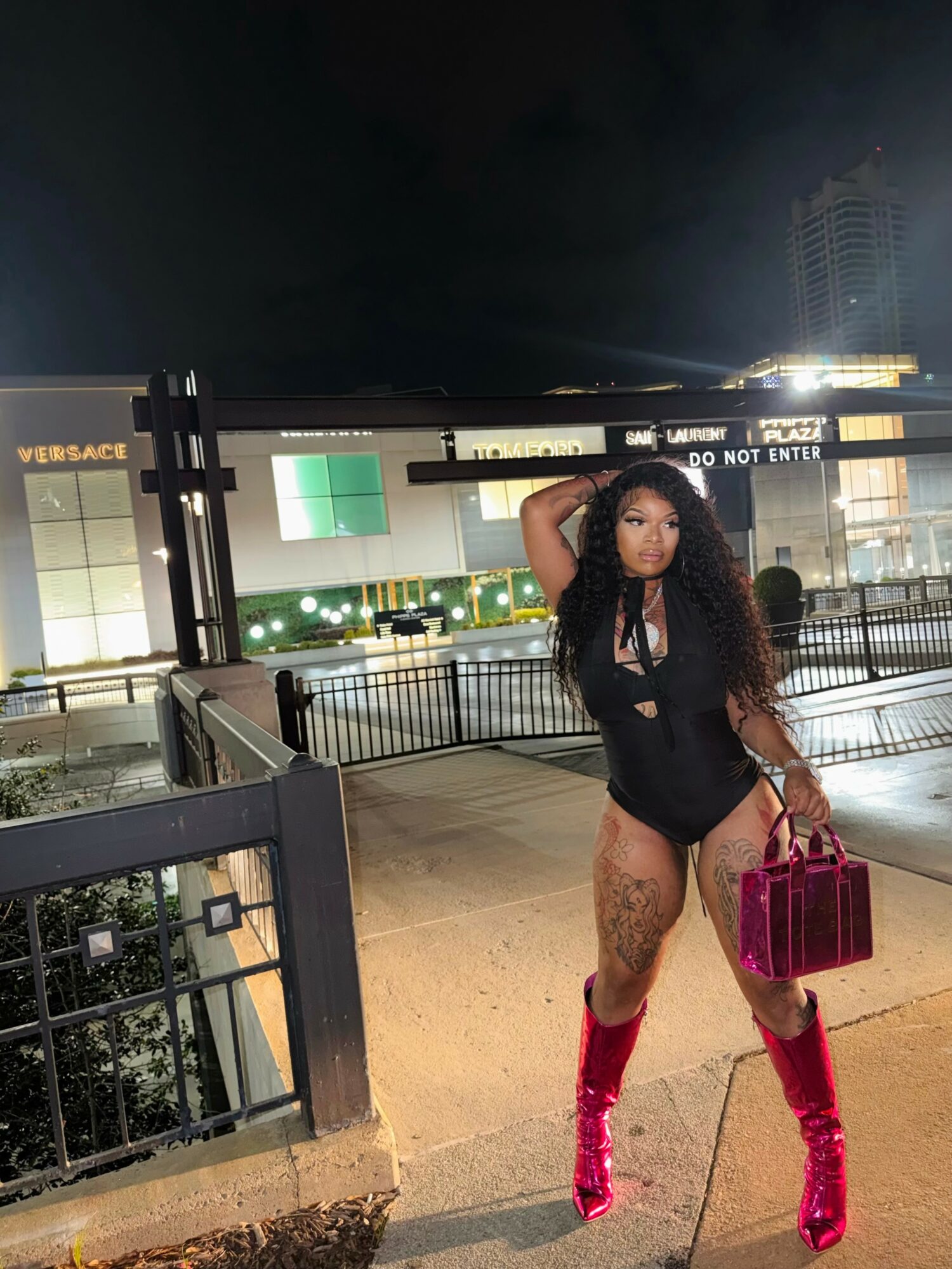 Woman with long hair in black outfit and red boots holding a red handbag on city street at night.