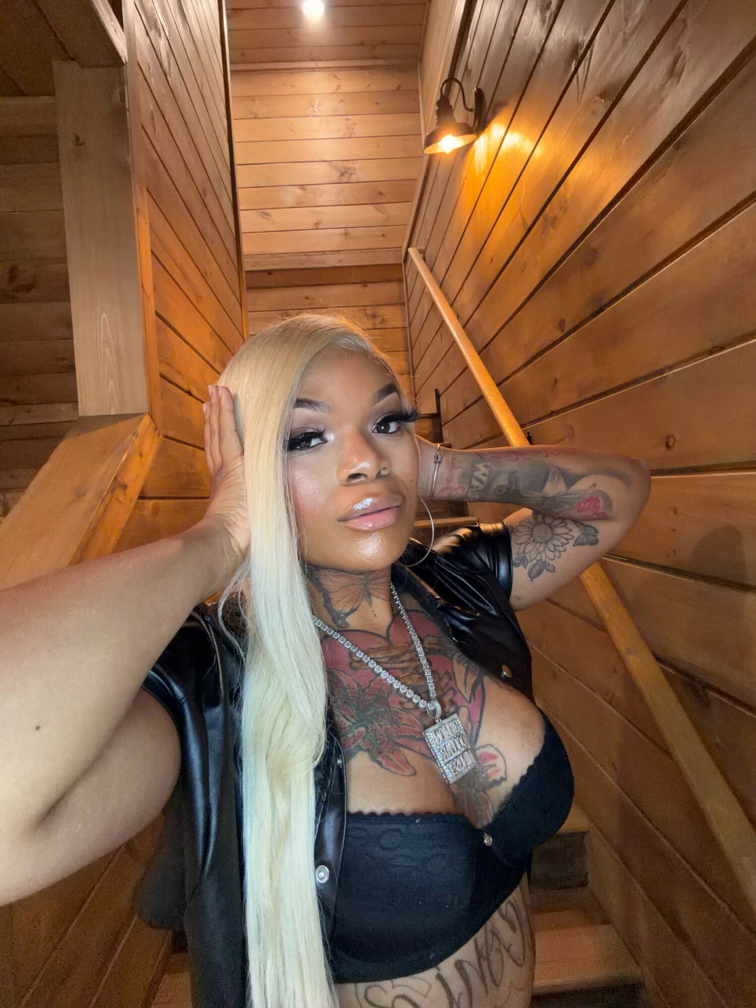 Woman with long blonde hair, tattoos, and black outfit, posing in wooden interior with warm lighting.