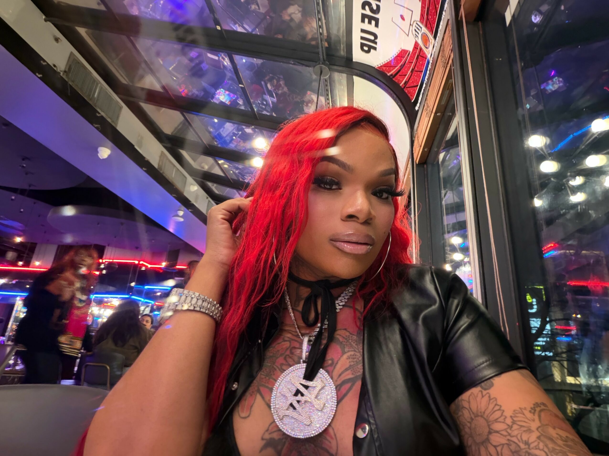 Woman with red hair and tattoos sitting indoors near a window with city lights outside.