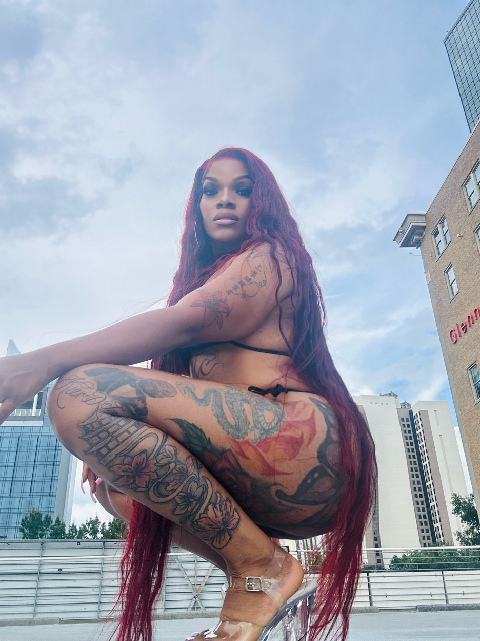 Woman with long red hair and tattoos crouching outdoors against city buildings and sky.