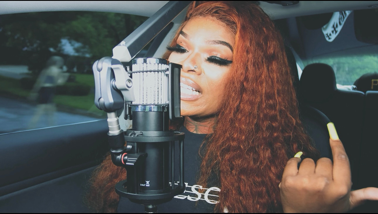 Woman with long curly hair speaking into a microphone inside a vehicle, with a blurred background outside the window.