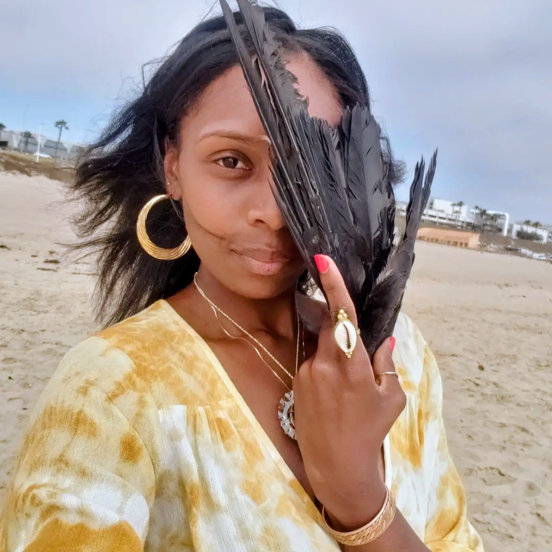 Woman holding black feathers in front of her face on a beach, wearing yellow and white clothing, gold jewelry.