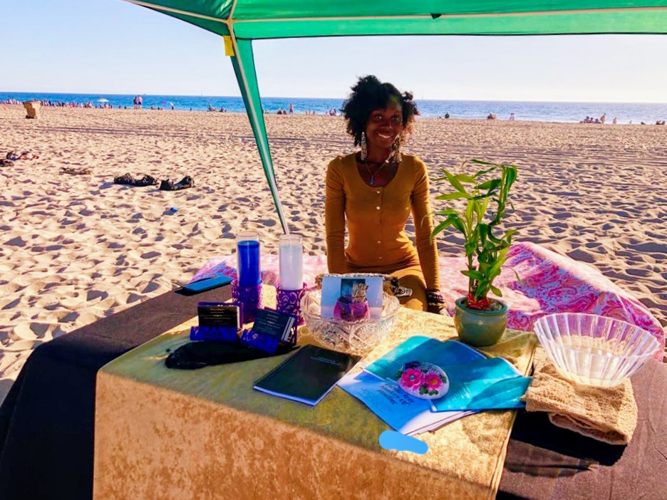 Woman sitting at a table on the beach under a green umbrella, with various items including a plant, books, and a tablet.