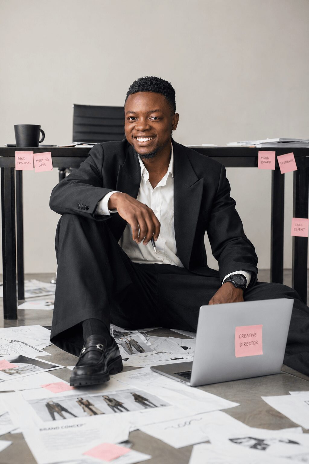 Young man in suit sitting on floor with laptop and papers, smiling in office setting.