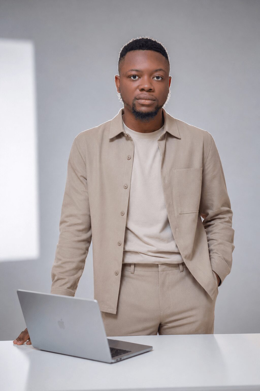 Young man standing behind a white table with a laptop, wearing beige shirt and pants, in a neutral room.