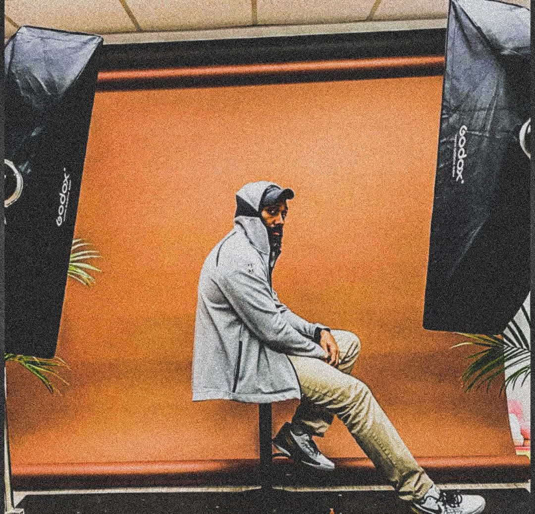 Person sitting on a stool in front of an orange backdrop, wearing a gray hoodie and beige pants.