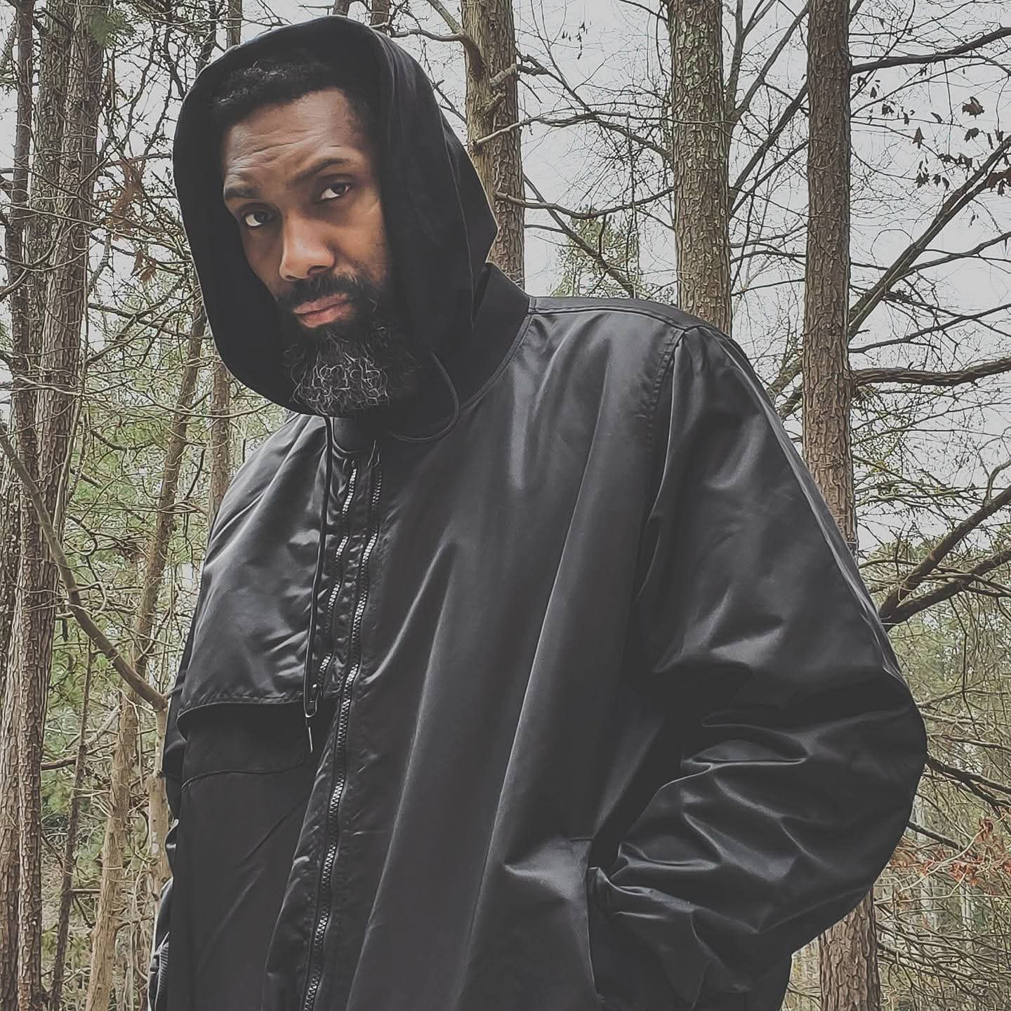 Man with a beard wearing a black hooded jacket outdoors in a wooded area.