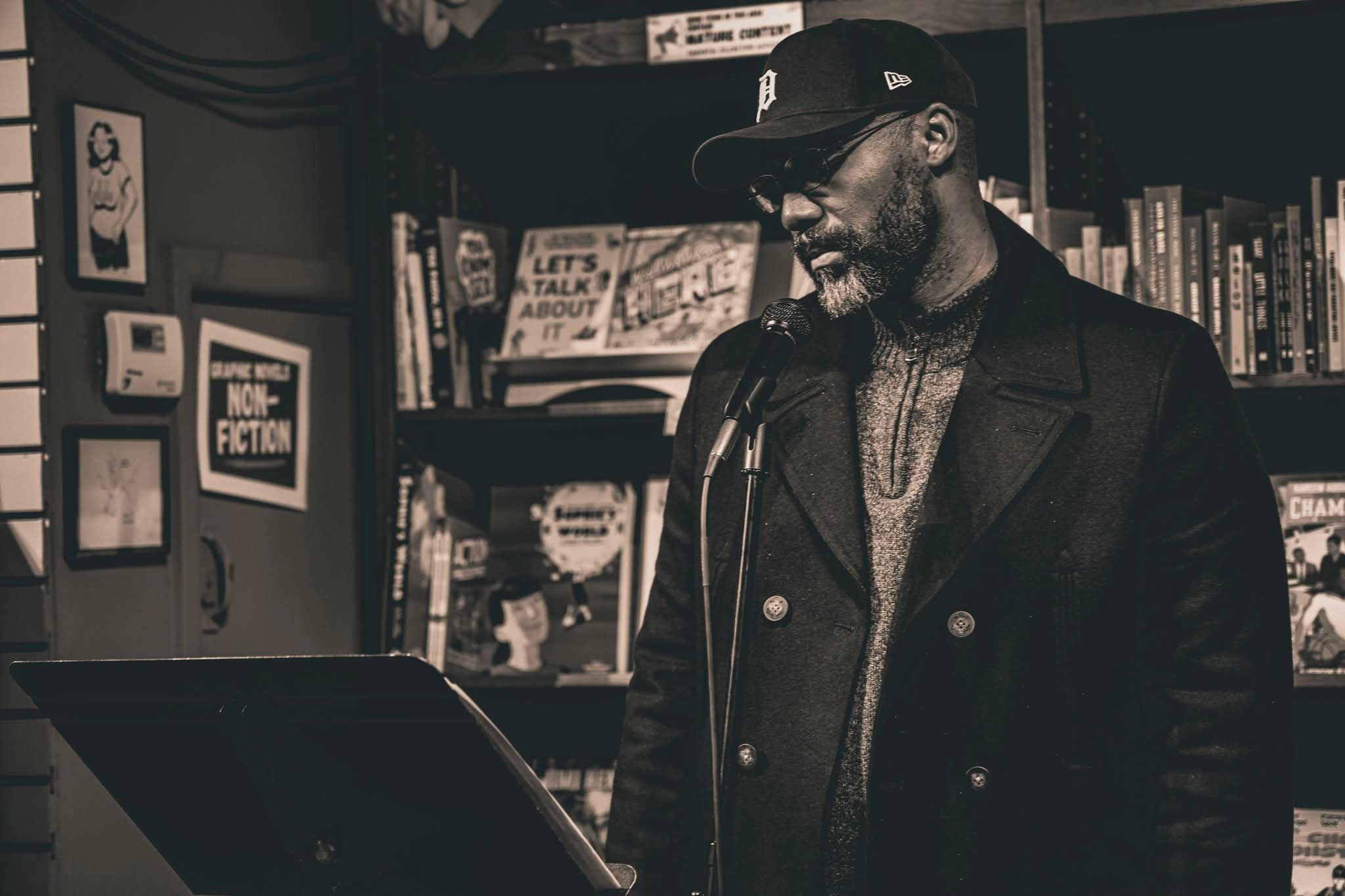 Man with glasses and beard wearing a cap and coat, looking at a book or tablet in a bookstore.