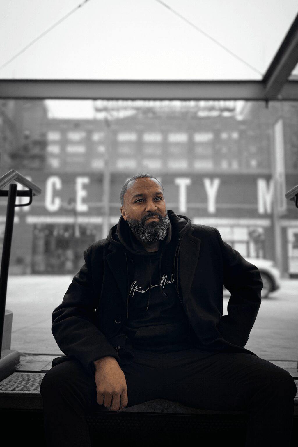 Man with beard sitting indoors near large window with cityscape background.