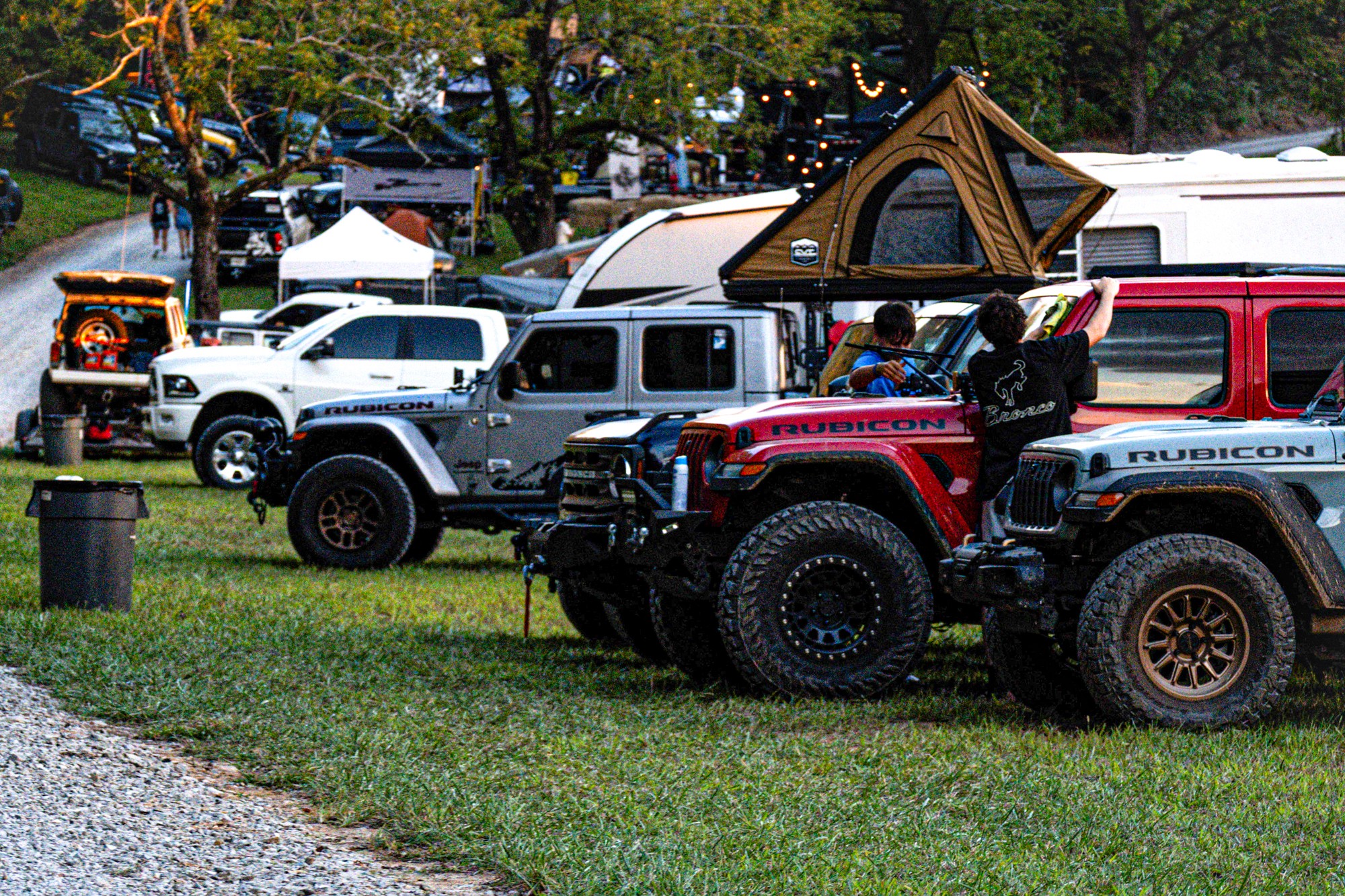 Line of parked trucks and SUVs on grass with trees and tents in background, some vehicles have rooftop tents.