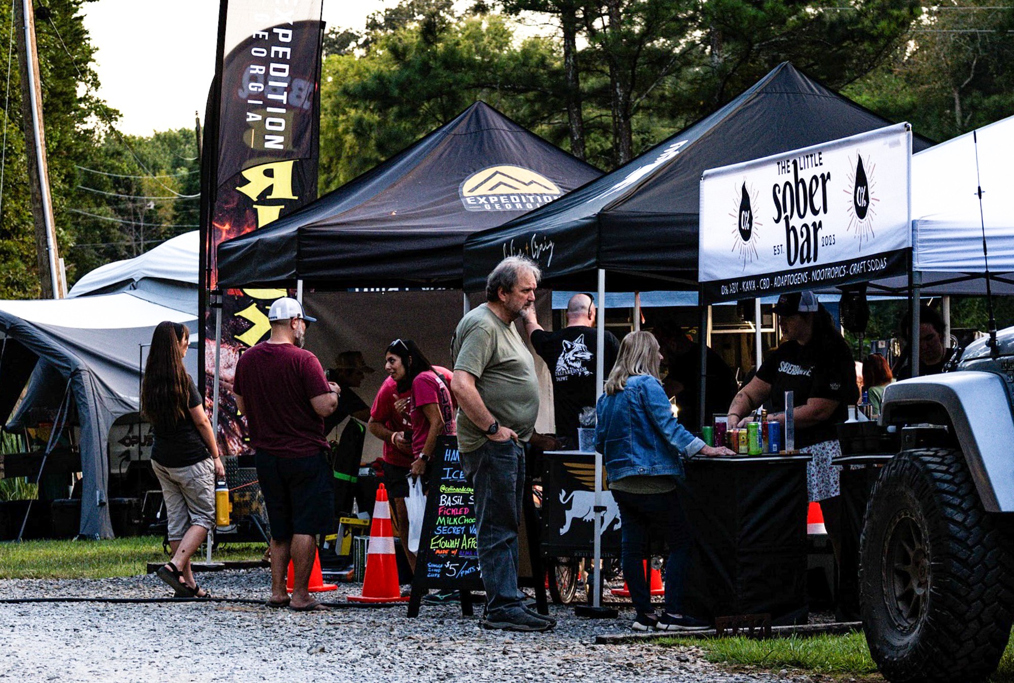 People gather around tents and a food truck at an outdoor event with trees in the background.