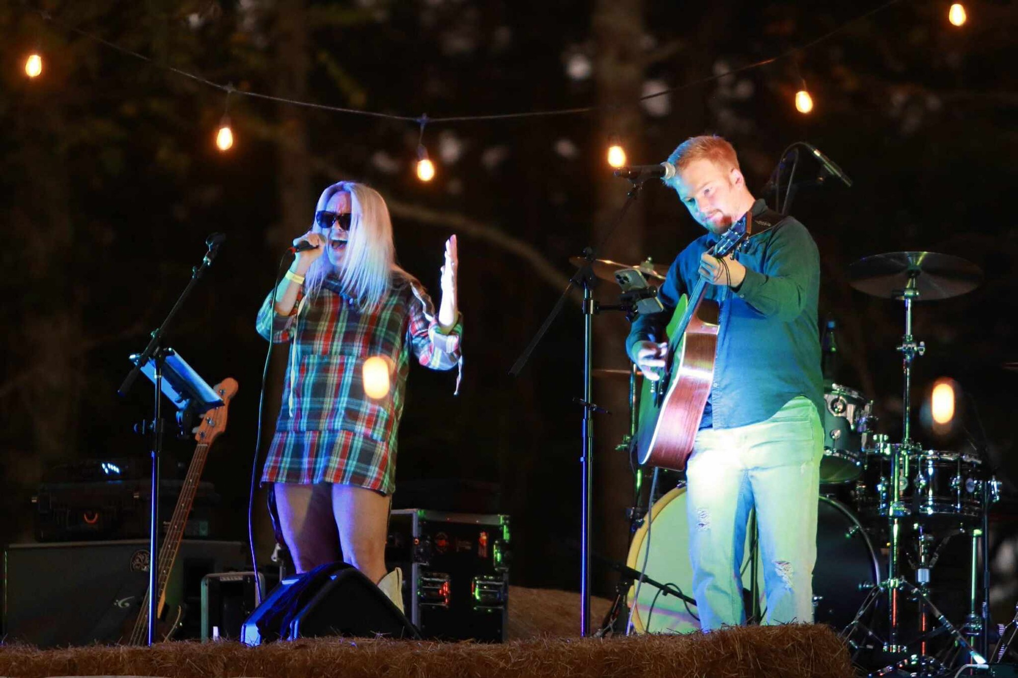 Two musicians perform on stage with string lights overhead, one singing into a microphone and the other playing guitar.