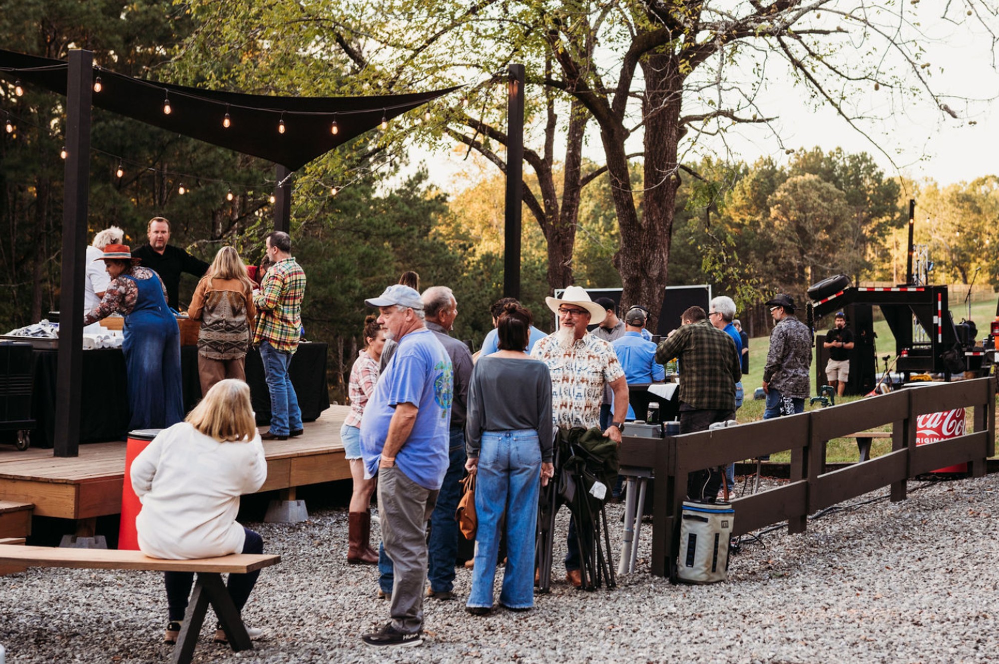 Group of people gathered outdoors near a stage with trees and string lights, some standing and some sitting.
