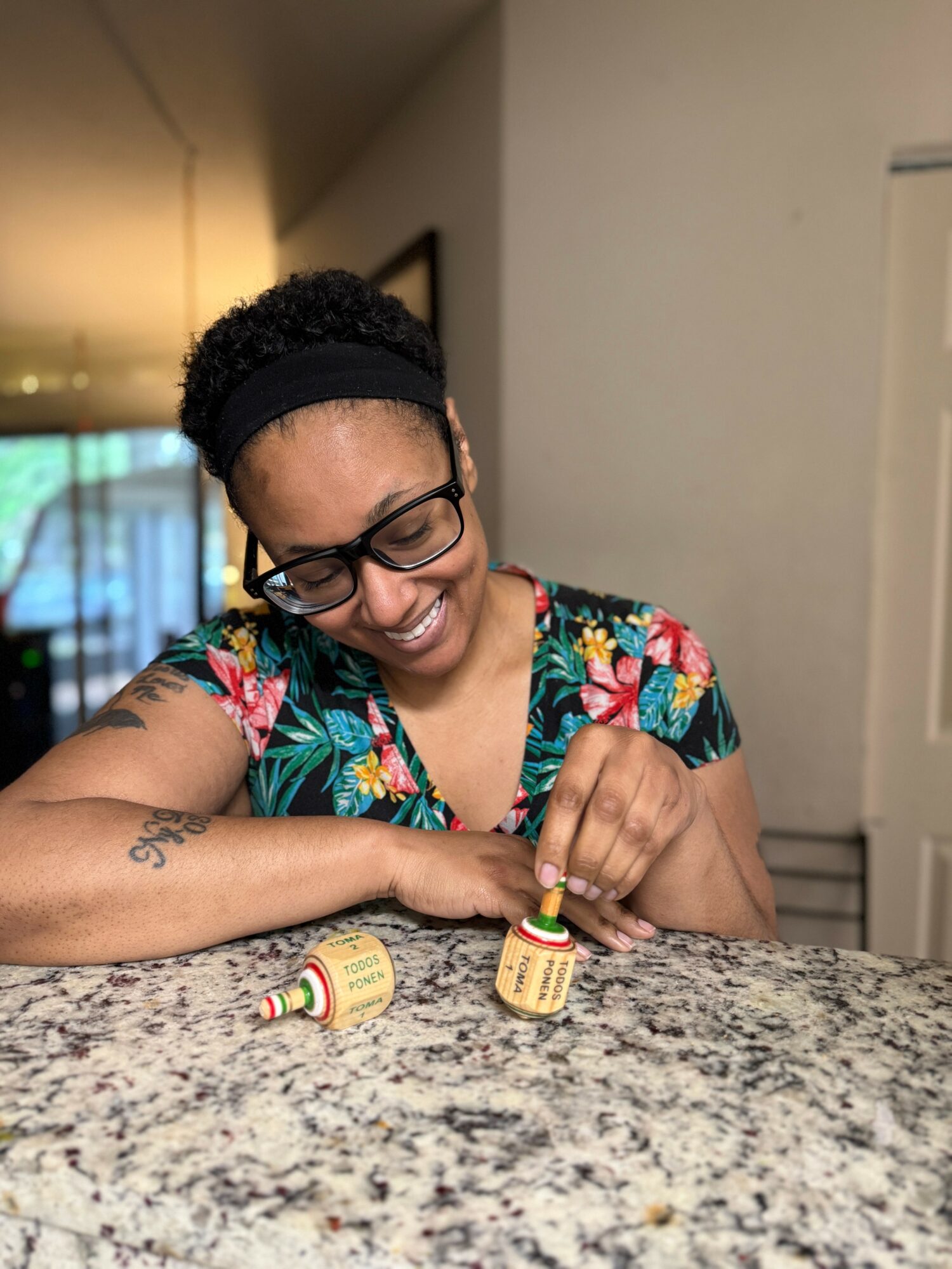 Woman with glasses and a floral shirt smiling while holding a small bottle on a kitchen counter.