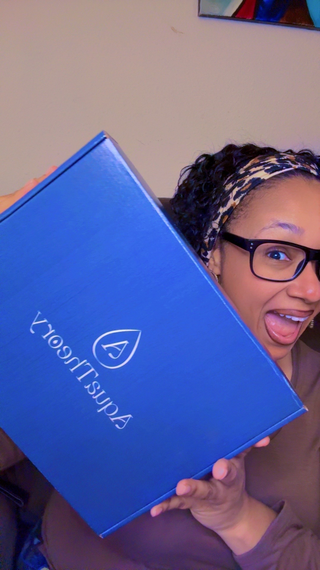 Person holding a blue box with a logo, smiling and wearing glasses and a headband.