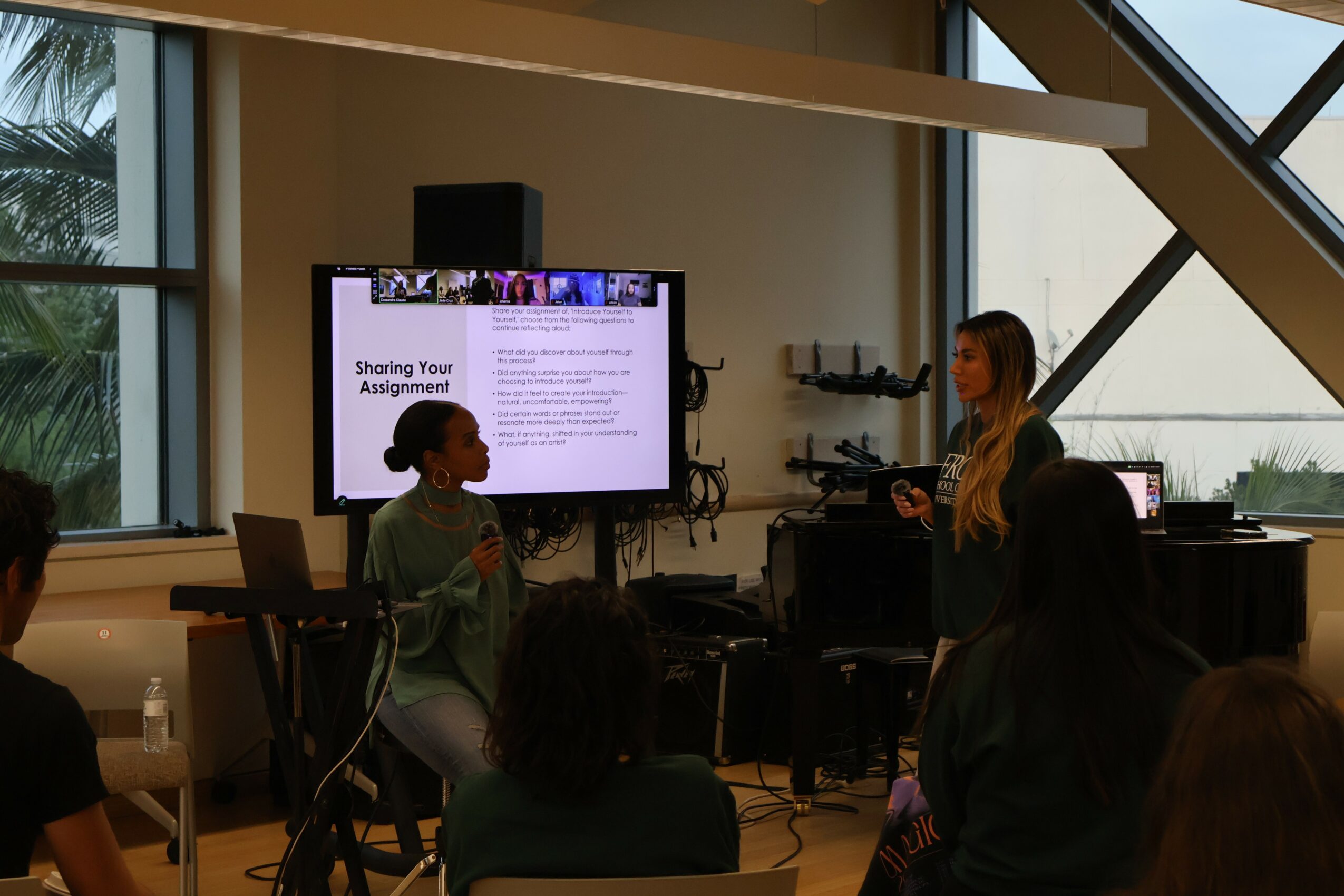 Two women speaking in a conference room with a large screen displaying a presentation slide, and audience members seated.
