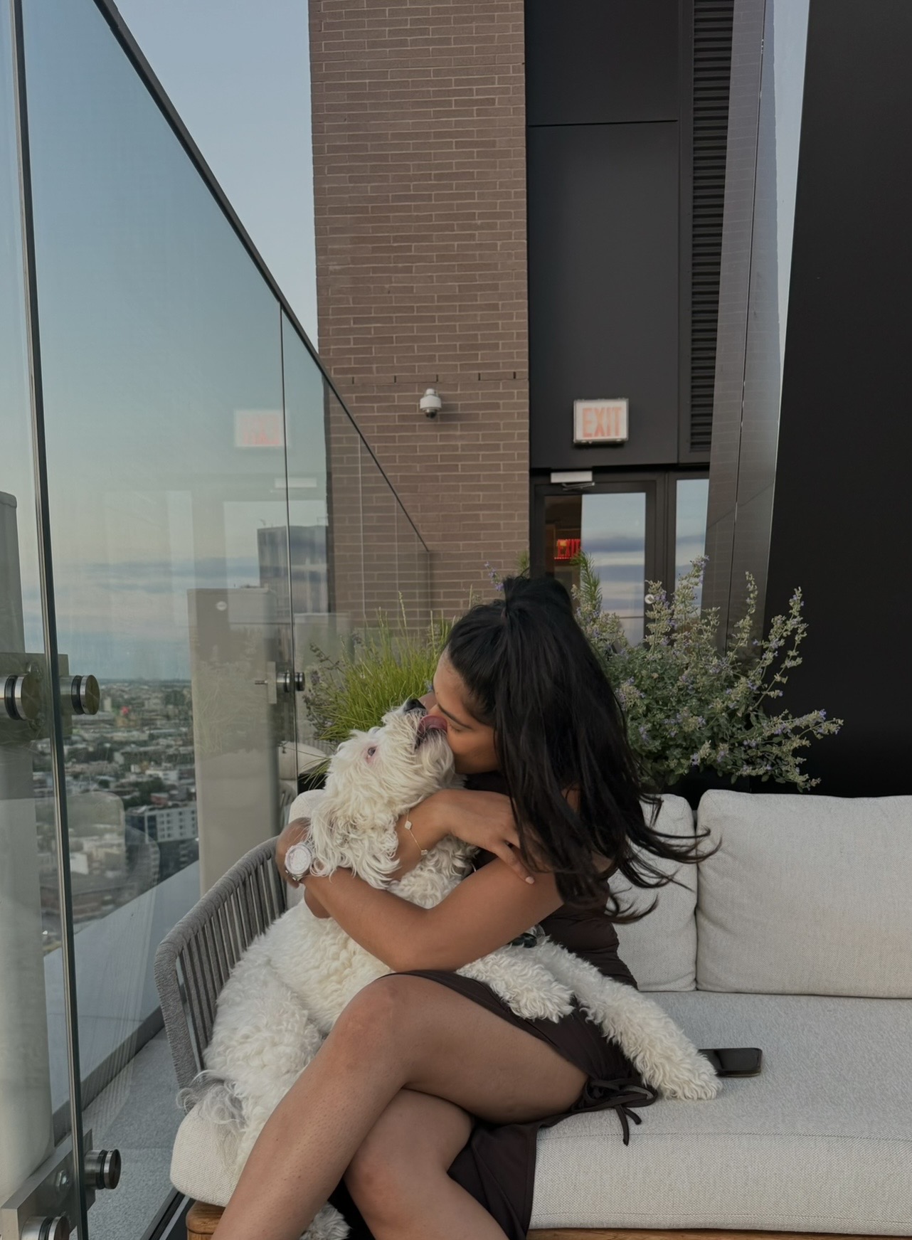 Woman with long dark hair sitting on a white sofa, hugging a fluffy white dog, with city view outside window.