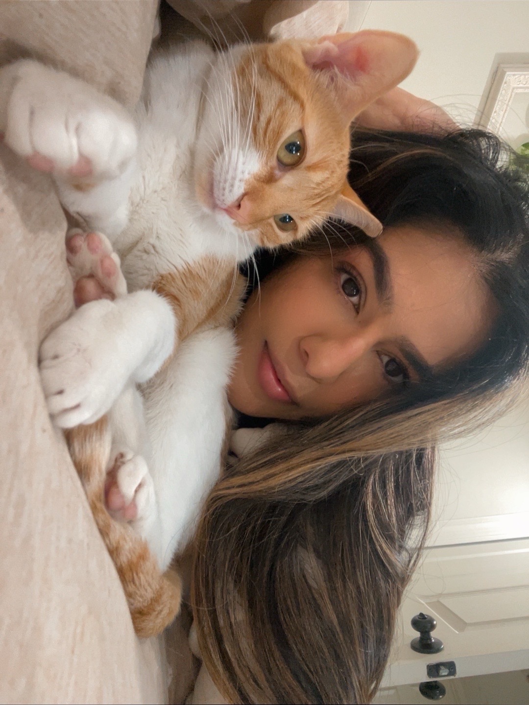 Woman lying down with an orange and white cat, both looking at the camera, indoors.