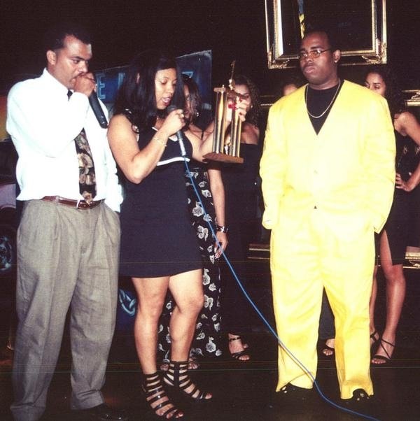 Three people standing on stage, one holding a trophy, with a dark background and framed mirror. The person on the right wears a yellow suit.