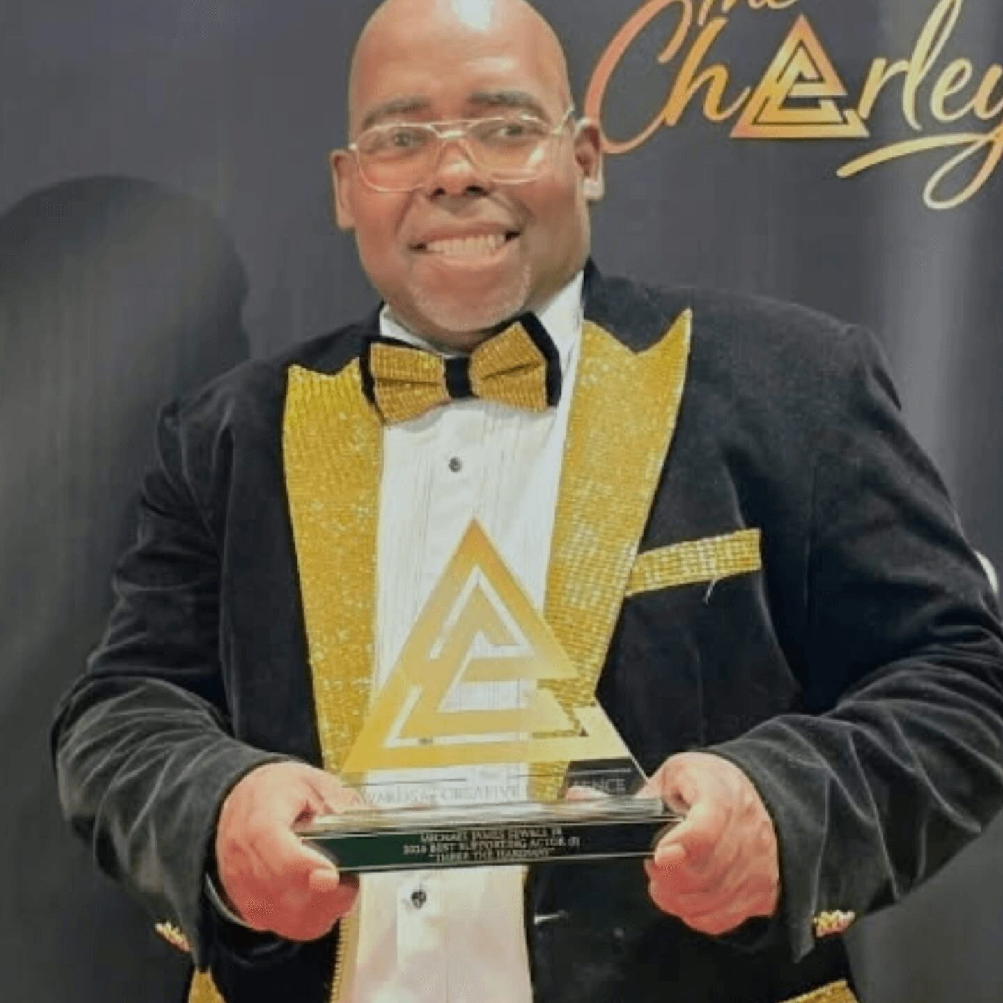Man in tuxedo holding an award, smiling, with a black background and gold lettering.