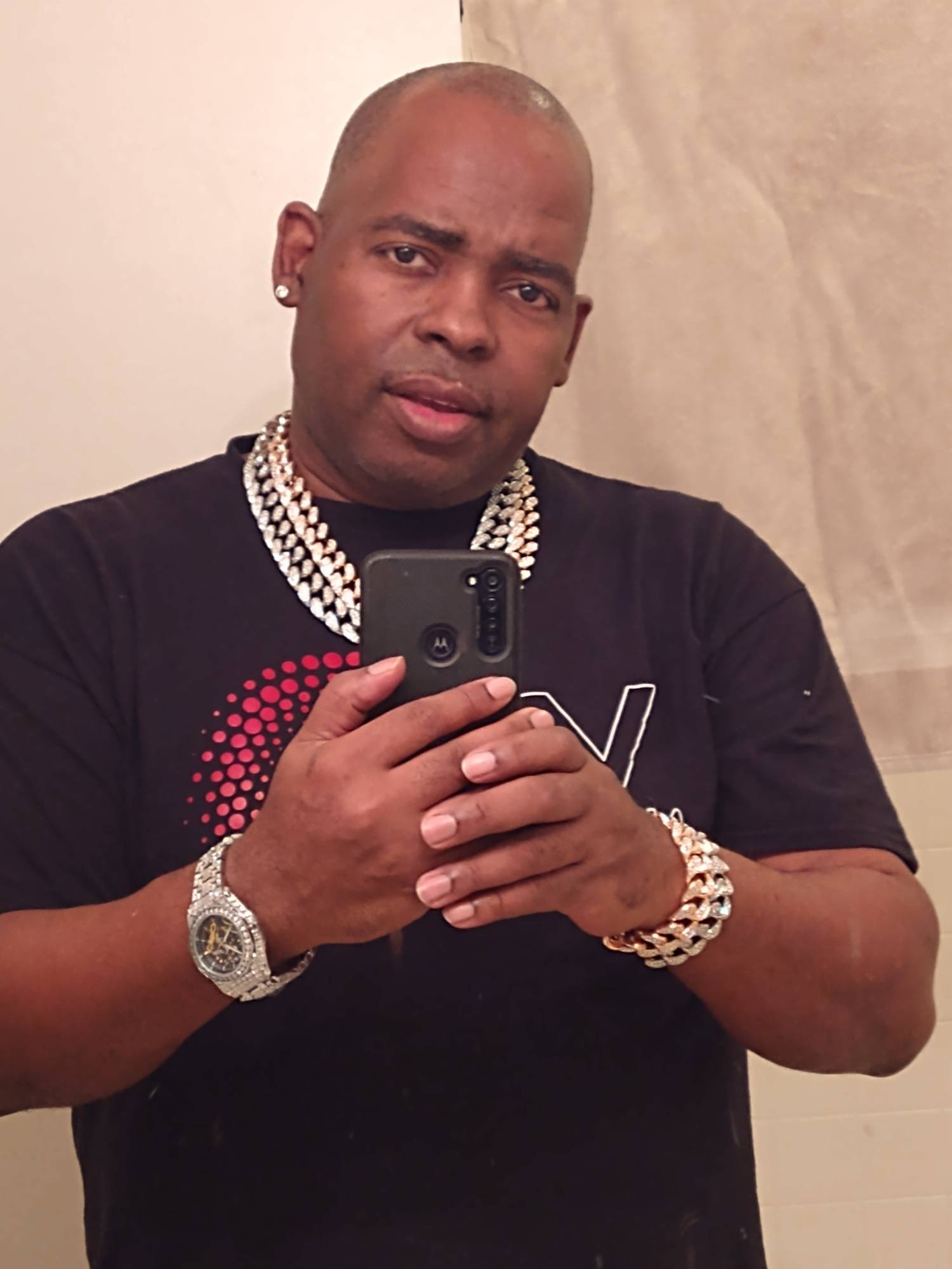 Man taking a selfie with a smartphone, wearing jewelry and a black T-shirt, in front of a beige wall.