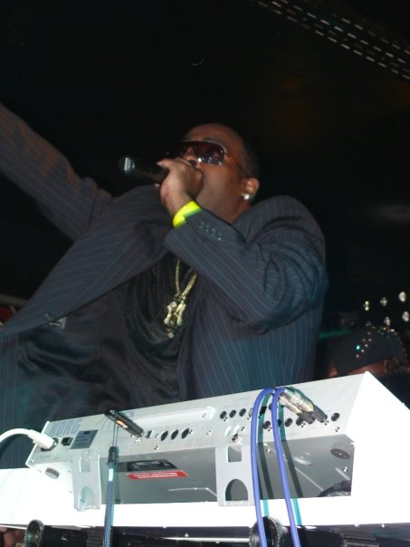 Person wearing sunglasses and a suit, holding a microphone, standing behind DJ equipment in a dark setting.