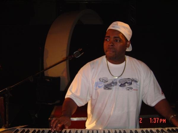 Person wearing a white cap and t-shirt, standing behind a keyboard on stage, with a microphone nearby.