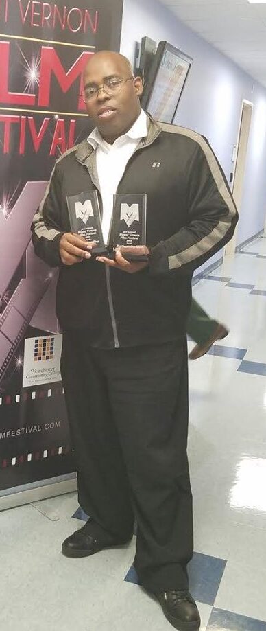 Man holding two awards in a hallway with a poster behind him.