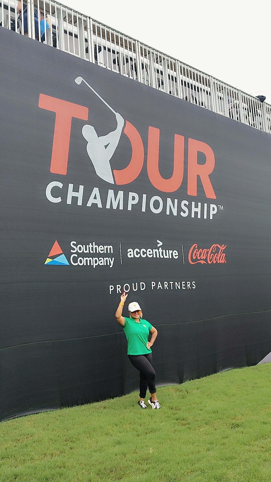 Person standing on grass in front of large black wall with TOUR CHAMPIONSHIP logo and sponsor logos.