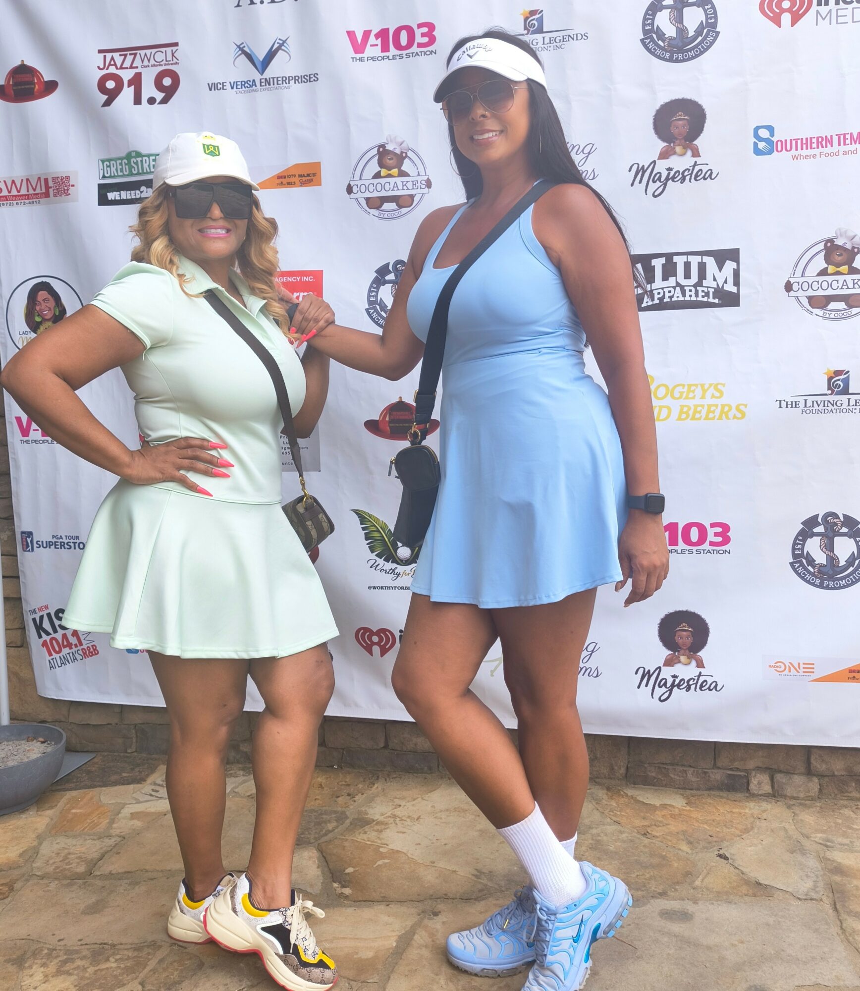Two women in tennis outfits and caps pose in front of a promotional backdrop.